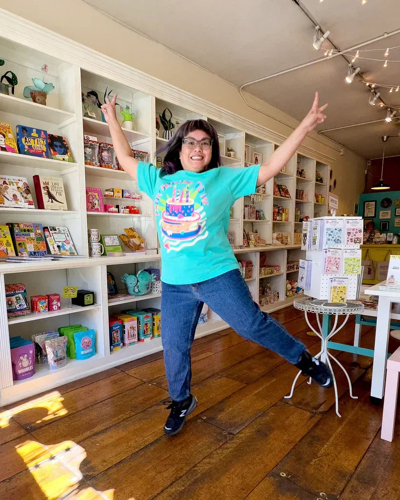 Image of Leanna, a middle-aged woman with black hair and glasses, jumping in the air inside her gift store.