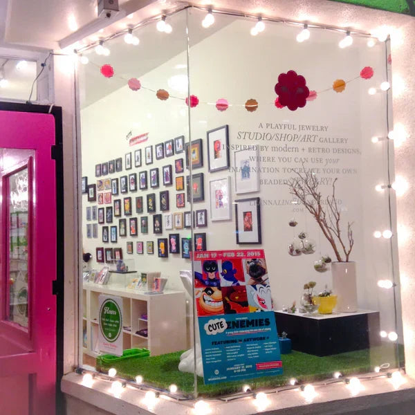 Photo of the outside window of Leanna Lin's Wonderland gift shop at night, looking in at the gallery wall, filled with many art pieces. Pink garland hangs in the window along with a string of lights.