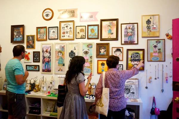 Photo of three people looking at the gallery wall for the Misha Lulu show. The wall features twenty-five plus art pieces including some tactile 3D pieces of animals, people, and more.