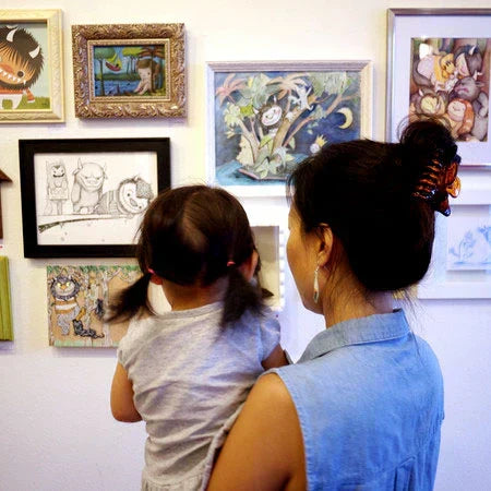 Photo of a woman holding a little girl while they both look at art hanging on a gallery wall. The art features characters from the book "Where the Wild Things Are". 