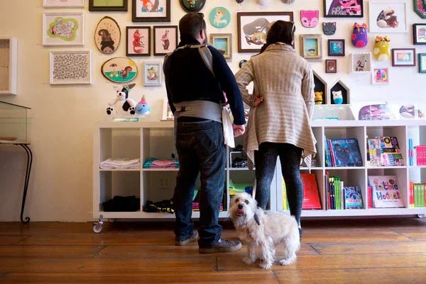 Photo of a man, woman, and their dog in front of the gallery wall for the "Paws" art show featuring what seems to be at least over thirty art pieces. The dog looks straight to camera in the photo.