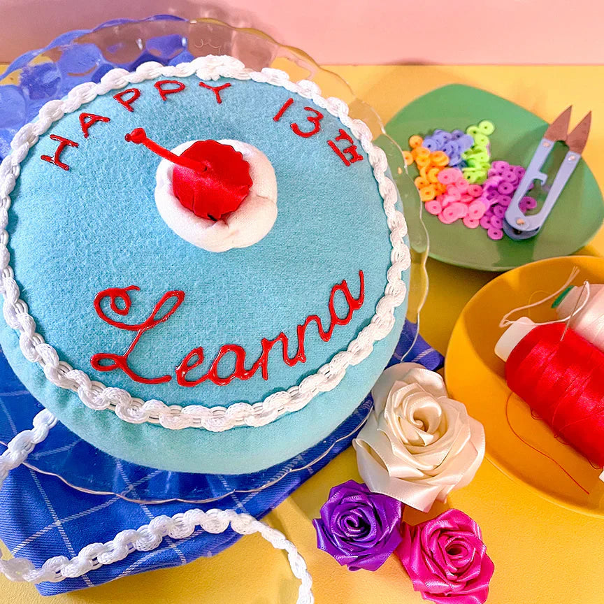 A blue felt cake with white ribbon icing, a cherry, and the words "Happy 13th Leanna" written on it. The cake is presented on a glass plate and is surrounded by crafting supplies.