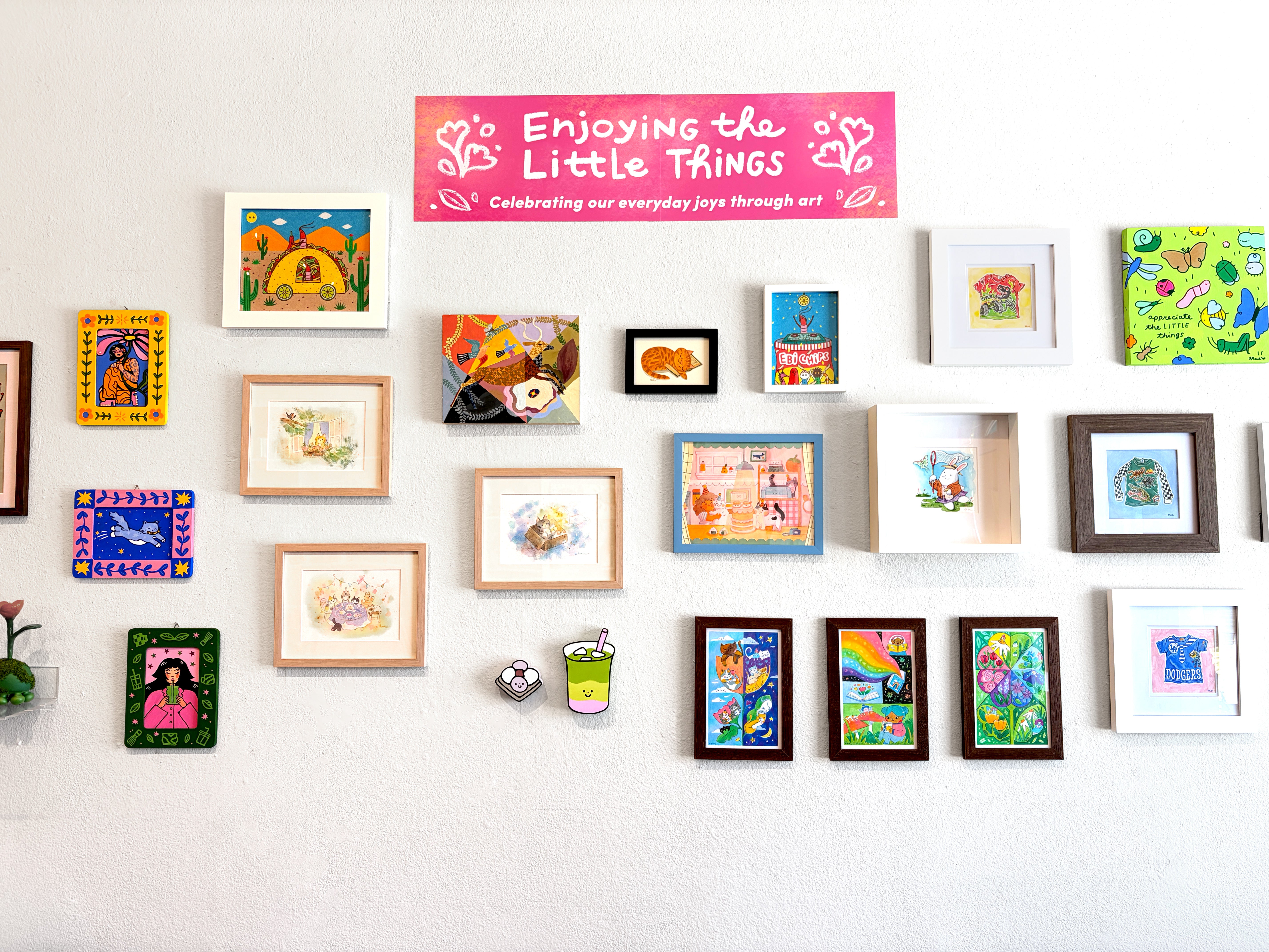 A gallery wall display featuring numerous colorful framed art prints, paintings, and small decorative pieces arranged on a white wall. A pink banner at the top reads 'Enjoying the Little Things, Celebrating our everyday joys through art'.