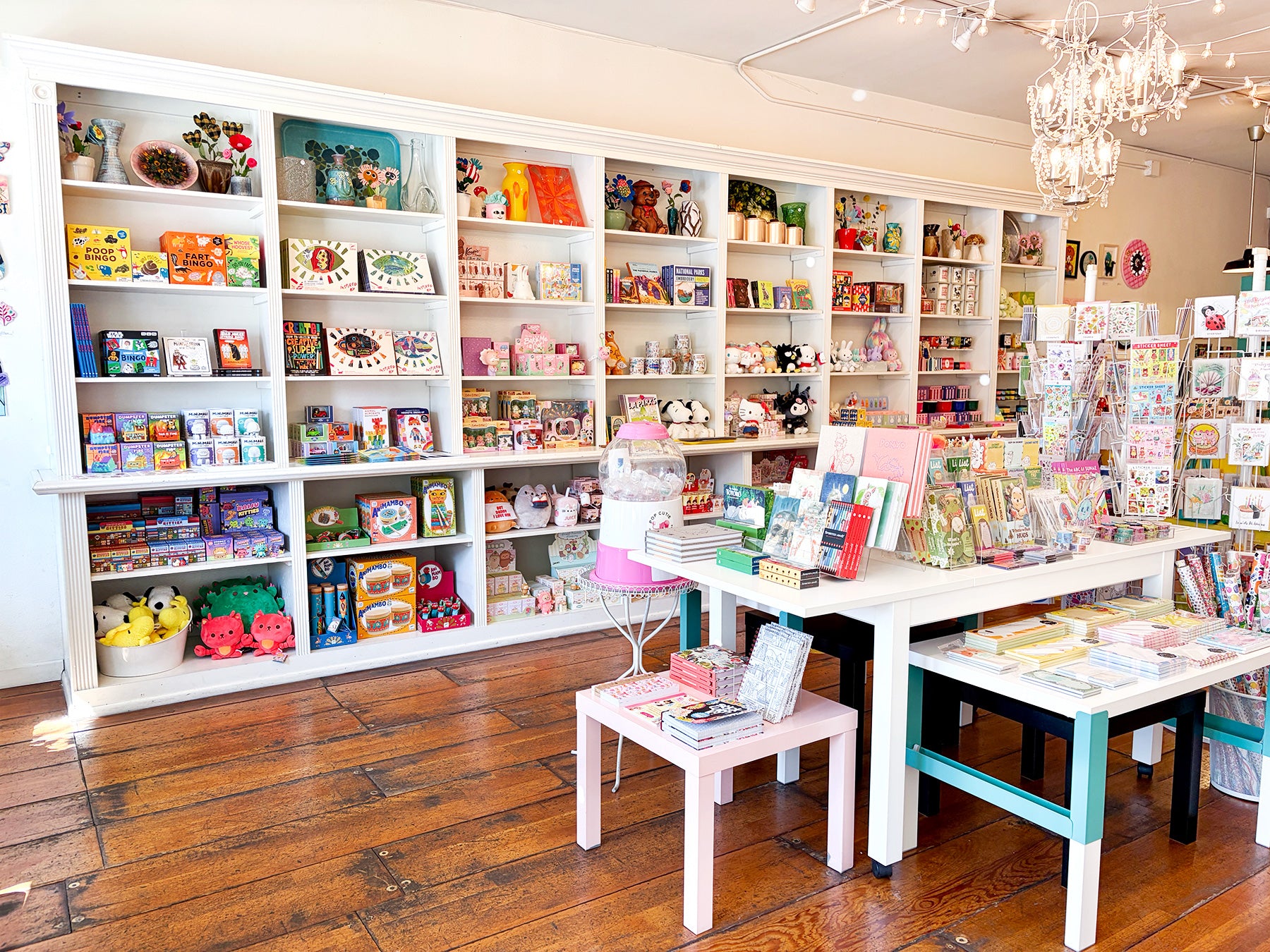 A bright, well-organized gift store interior with white shelving units filled with colorful books, toys, and gift items, alongside small display tables on a polished wood floor and a small chandelier.