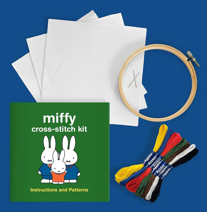 Miffy cross-stitch kit laid out on a blue background, including white fabric squares, a wooden embroidery hoop, a green booklet labeled &