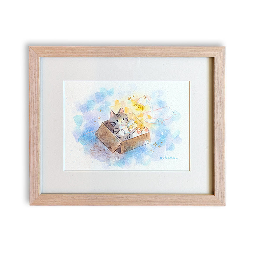 A framed watercolor art print featuring a small gray kitten sitting and reading in a brown box with a reading light, set in a light natural wood frame with a white mat.