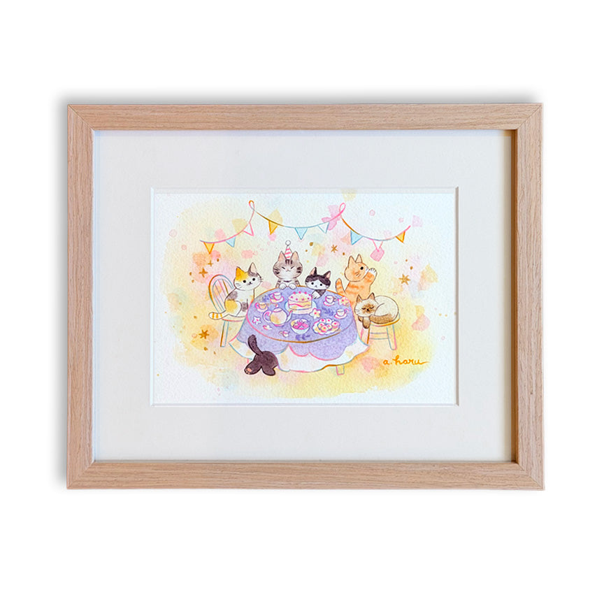A framed watercolor art print featuring cute five cartoon cats gathered around a purple table with a cake and one cat peeking from under the table, set in a light natural wood frame with a white mat on a white background.