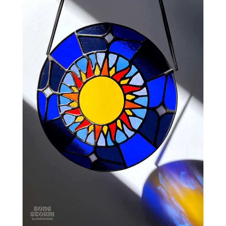 A bright circular stained glass suncatcher featuring a deep blue glass and yellow and orange sun design, hanging by a black cloth string near a window with bright sunlight and shadows.  