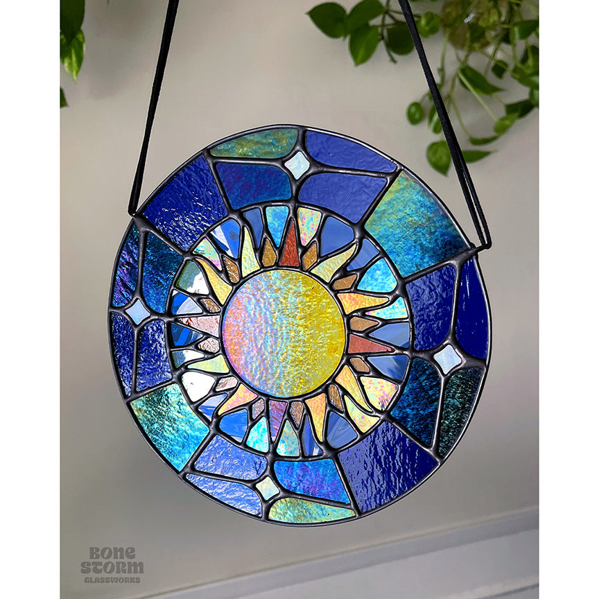 A bright circular stained glass suncatcher featuring a deep blue glass and yellow and orange sun design, hanging by a black cloth string, slightly muted with no sun reflected on it.