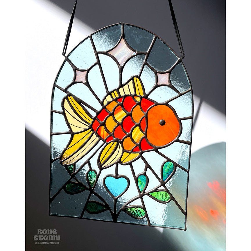 A bright stained glass suncatcher featuring an orange goldfish design, hanging by a black cloth string near a window with bright sunlight and shadows. 