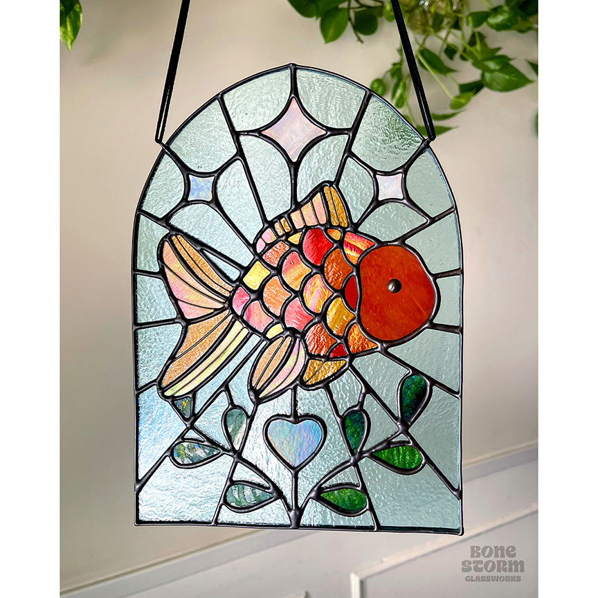 A bright stained glass suncatcher featuring an orange goldfish design, hanging by a black cloth string, slightly muted with no sun reflected on it.