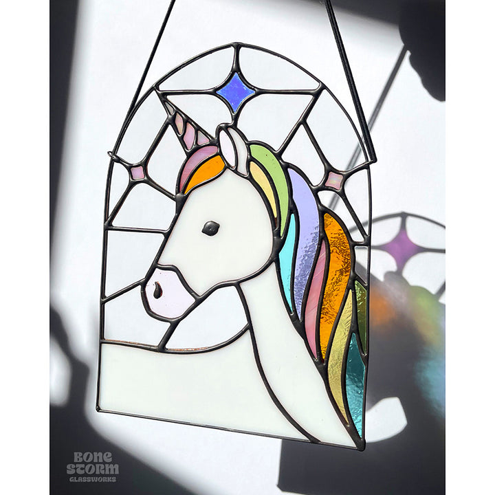 A stained glass suncatcher with a white and pastel rainbow unicorn design. Hanging by a black cloth string near a window with bright sunlight and shadows. 