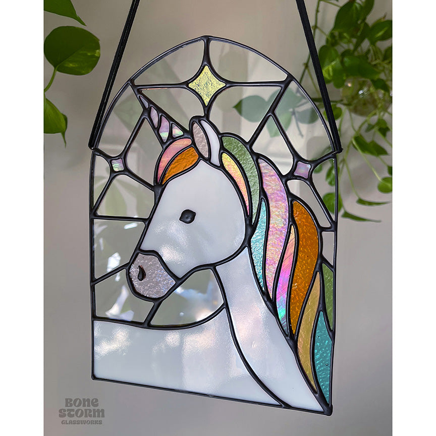 A stained glass suncatcher with a white and pastel rainbow unicorn design. Hanging by a black cloth string, slighted muted with no sun reflected on it.