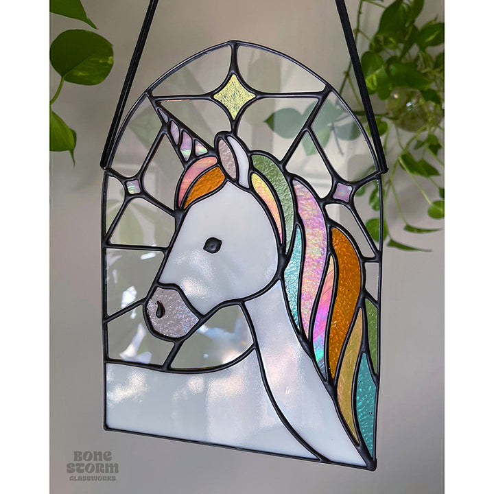 A stained glass suncatcher with a white and pastel rainbow unicorn design. Hanging by a black cloth string, slighted muted with no sun reflected on it.