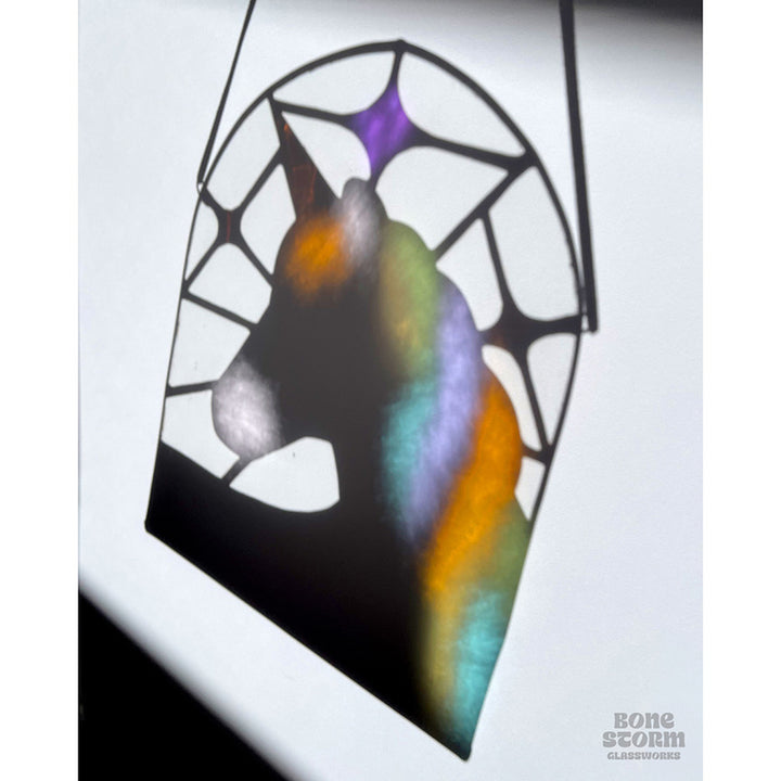 Colorful reflection on a wall of a stained glass suncatcher with a white and pastel rainbow unicorn design. Design is such that the shadow on the wall looks like that of a unicorn in profile. 