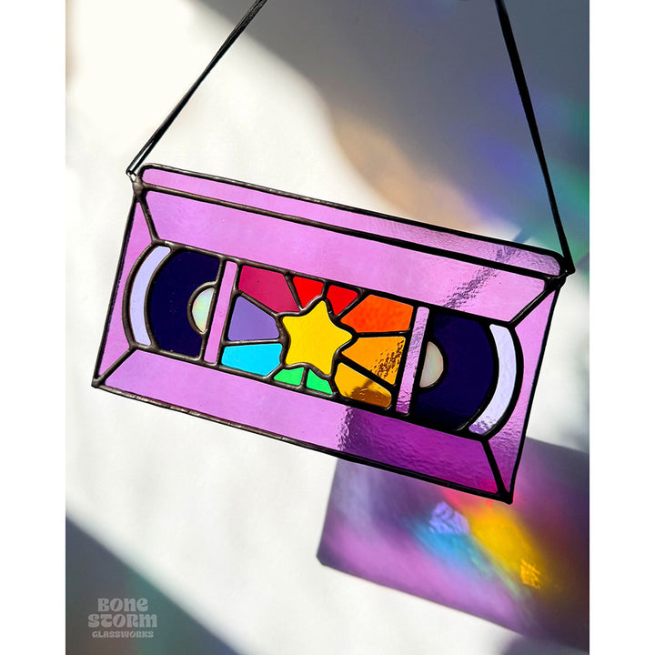 A bright stained glass suncatcher shaped and designed like a VHS tape. The tape is purple in color with a yellow star and rainbow colored glass panes in the middle. Hanging by a black cloth string near a window with bright sunlight and shadows. 