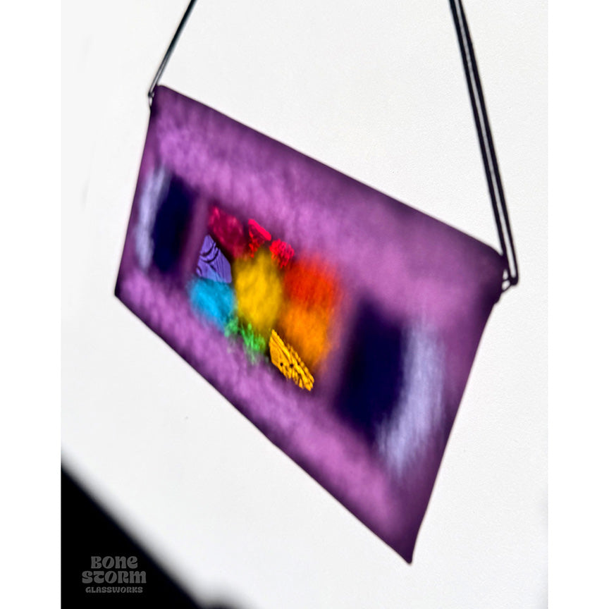 Colorful reflection on wall of a bright stained glass suncatcher shaped and designed like a VHS tape. The tape is purple in color with a yellow star and rainbow colored glass panes in the middle. 