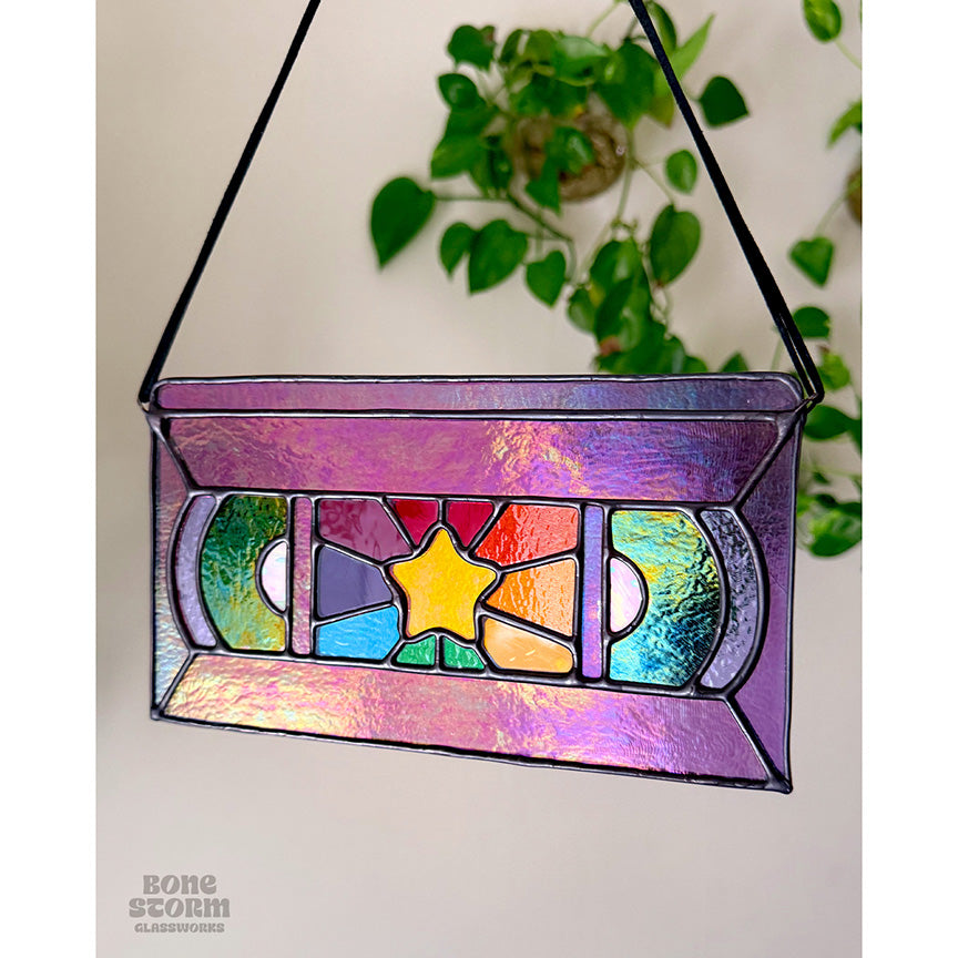 A stained glass suncatcher shaped and designed like a VHS tape. The tape is purple in color with a yellow star and rainbow colored glass panes in the middle. Hanging by a black cloth string, slightly muted with no sun reflected on it.
