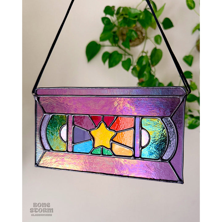 A stained glass suncatcher shaped and designed like a VHS tape. The tape is purple in color with a yellow star and rainbow colored glass panes in the middle. Hanging by a black cloth string, slightly muted with no sun reflected on it.