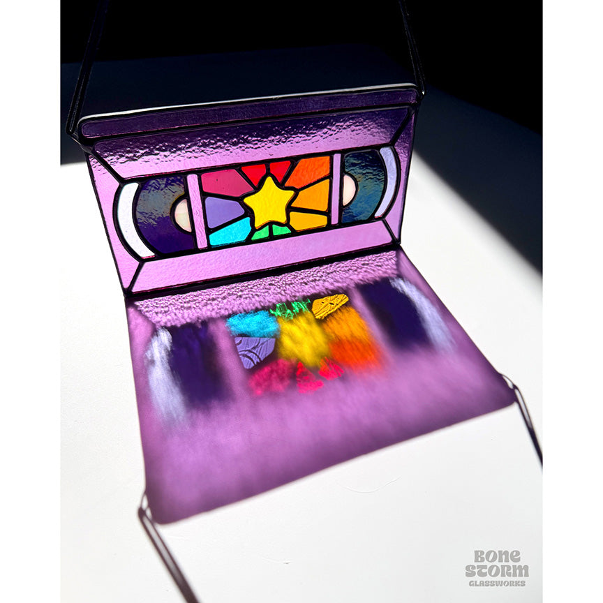 Colorful reflection on the ground, of a bright stained glass suncatcher shaped and designed like a VHS tape. The tape is purple in color with a yellow star and rainbow colored glass panes in the middle. 