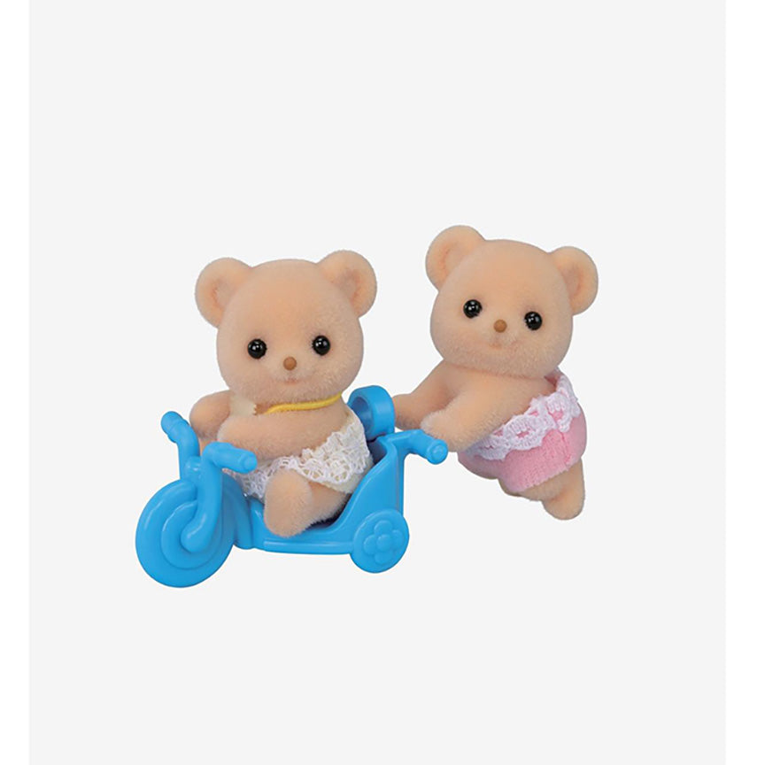 A pair of small teddy bear toy figures with soft peach flocked fur, one sitting in a blue tricycle and one pushing it, shown on a plain light gray background.