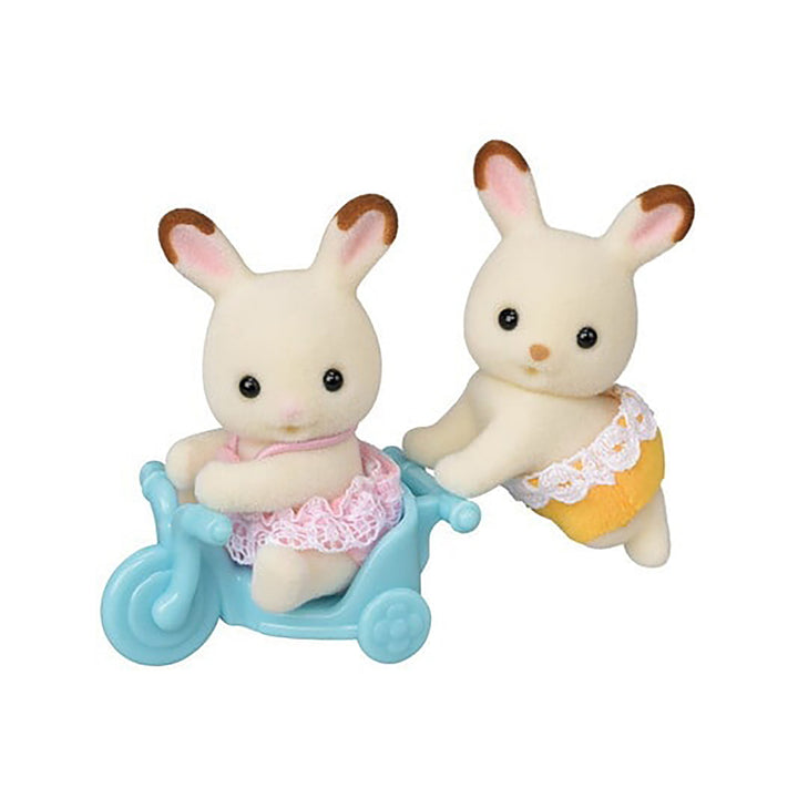 Two felted bunny rabbit toys with brown-tipped ears. One is crawling wearing yellow shorts while the other is in a blue plastic tricycle wearing a pink dress. 