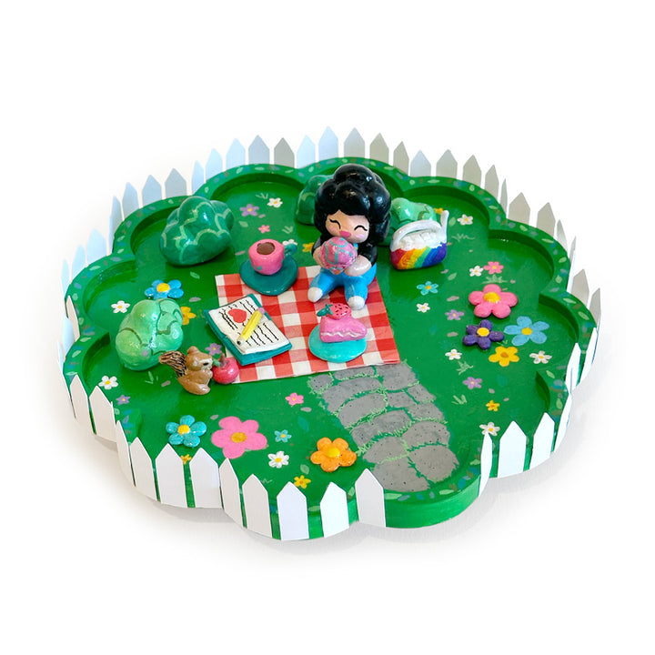 A colorful miniature sculpture depicting a picnic scene on a green grassy lawn with a white picket fence border, featuring a small figurine, flowers, rocks, and accessories.
