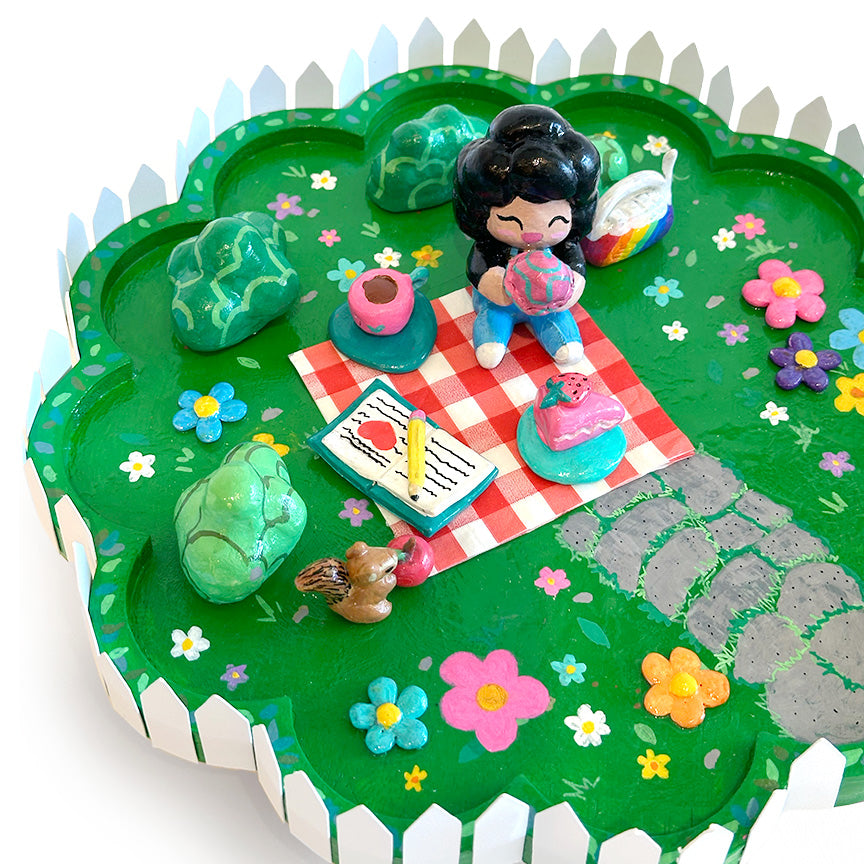 A colorful miniature sculpture depicting a picnic scene on a green grassy lawn with a white picket fence border, featuring a small figurine, flowers, rocks, and accessories.