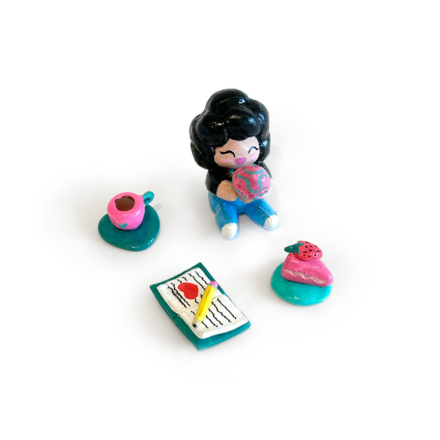 A small colorful figurine of a girl with black hair sitting and holding a pink ice cream cone, surrounded by three miniature accessories: a pink cup on a teal saucer, a pink slice of cake on a teal base, and a notebook with a yellow pencil.