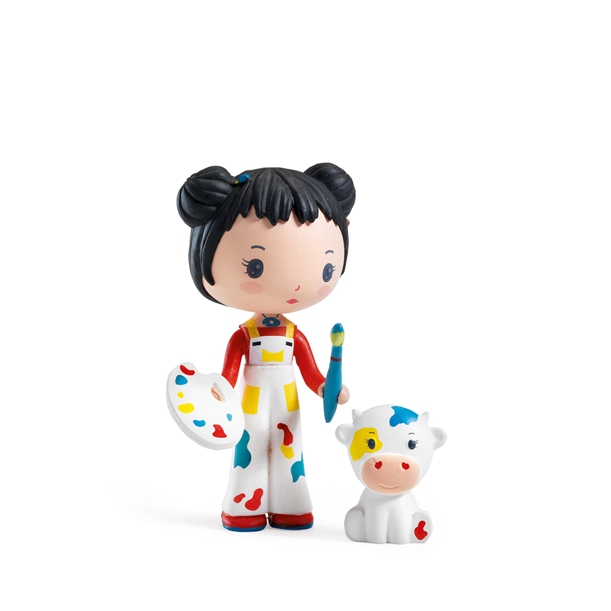 A toy figurine of a pale-skinned girl with black hair in twin buns, wearing white overalls with colorful paint splatters, holding a paintbrush and an artist palette next to a small white cow with painted spots, displayed on a white background.