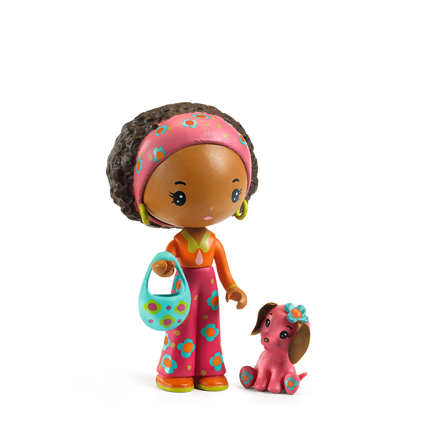 A toy figurine with clear brown skin, curly black hair, a pink patterned headband, and an orange top with colorful floral pants. She is holding a turquoise patterned handbag and stands next to a small pink dog figure with a blue flower headband. Figurines are shown on a white background. 