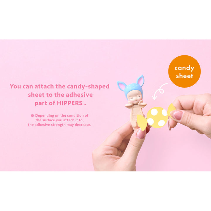 Hands attaching a yellow candy-shaped card with white dots to a small "sonny angel" figure with blue deer ears. Shown on a pink background with instructional text that reads: "You can attach the candy-shaped sheet to the adhesive part of Hippers."