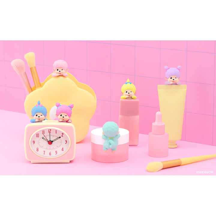 A pastel pink and yellow cosmetic-themed miniature display featuring small character figurines placed on and around makeup items, including a clock, brushes, cream tubes, a bottle, and a tube, set against a pink tiled background.