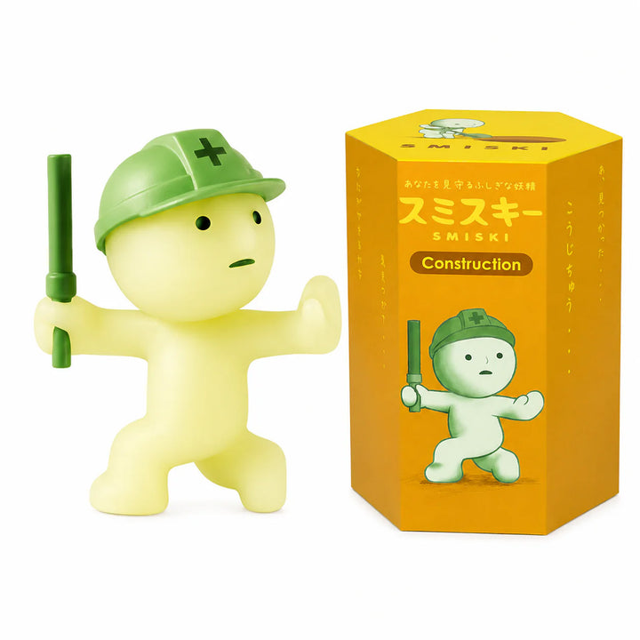 A pale yellow construction-themed figurine wearing a green hard hat and holding a green baton, shown next to its orange SMISKI product box on a white background.
