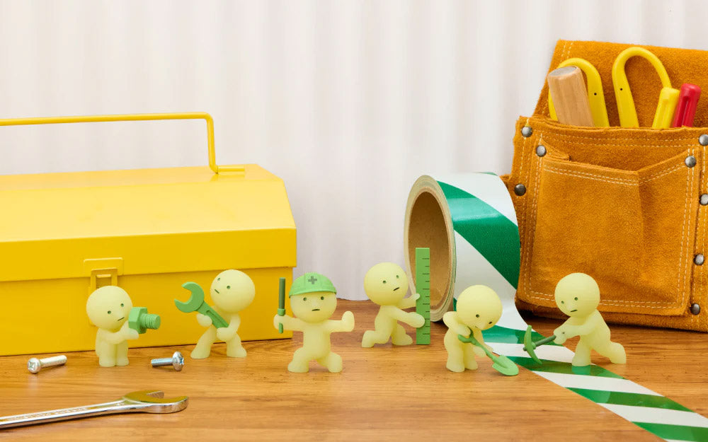 Toy figures of construction workers with tools on a wooden surface, including a yellow toolbox and orange tool bag.