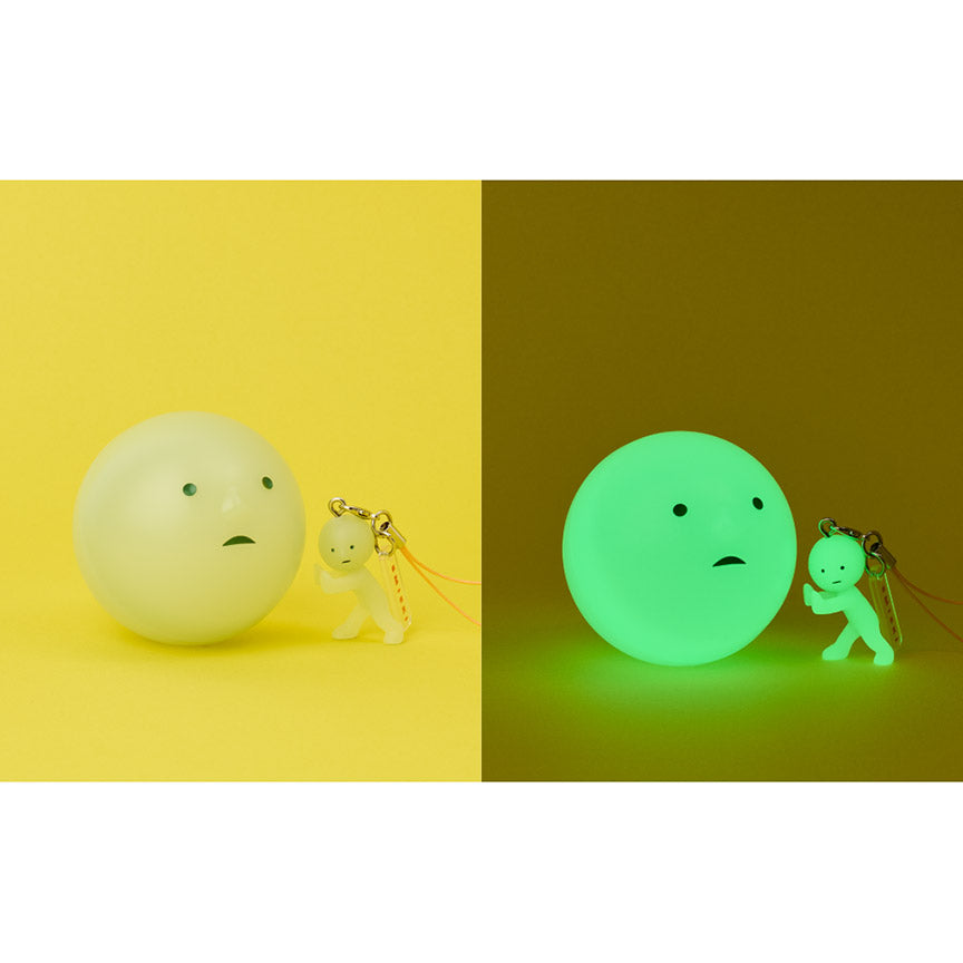 Two images showing a Smiski ball capsule and keychain on a yellow background, with the second picture depicting the same display glowing in the dark.