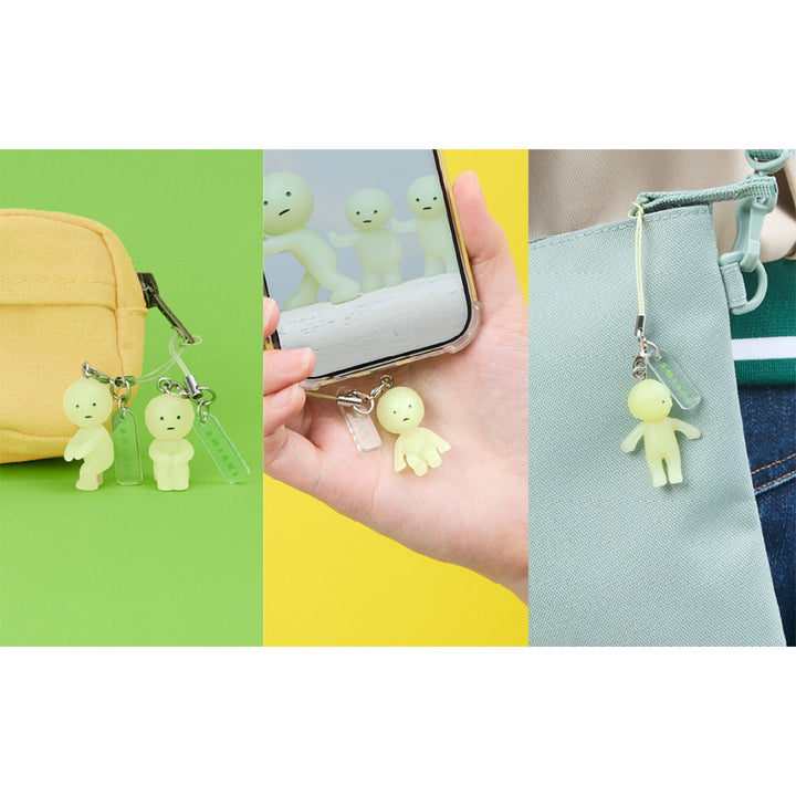Three examples of Smiski keychain uses; two displayed on the zipper of a pencil case, one on the bottom of a phone case, and one hung from a tote bag strap.