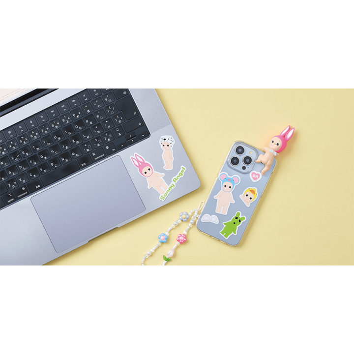 A clear phone case with cute cartoon bunny and character stickers, shown on a phone next to a silver laptop on a pale yellow background. The case has a dangling beaded strap with small pastel charms.