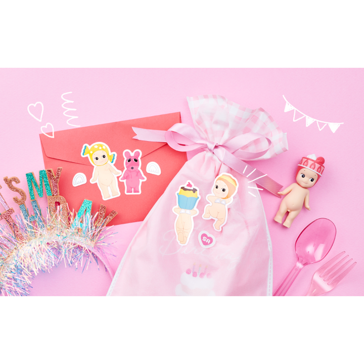 Pink birthday themed party accessories arranged on a pink background, including a sheer pink gift bag with baby character stickers, a red envelope, pink party utensils, a glittery tinsel crown, and a small Sonny Angel figurine.