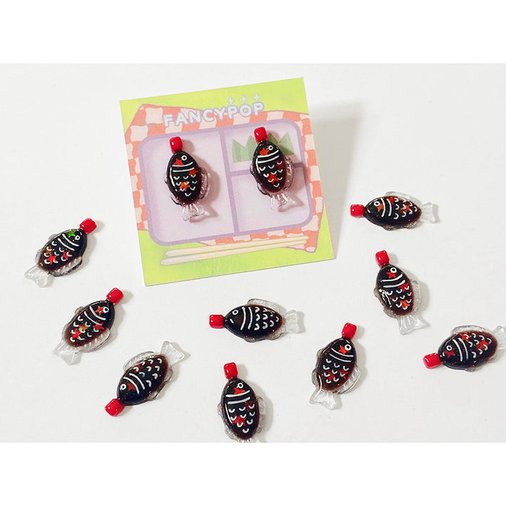Several pairs of fish-shaped soy sauce packet stud earrings, one pair are packaged on a bento box themed cardstock that says &