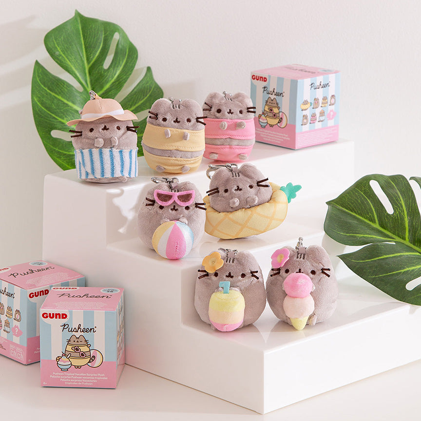 Seven Pusheen plush toys in different summer-themed outfits arranged on white display steps, with two pink GUND Pusheen boxes nearby and green monstera leaves in the background.
