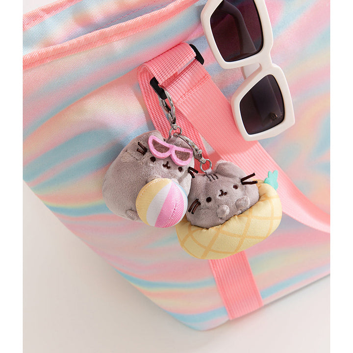 A pastel rainbow striped tote bag with a pink strap, decorated with two tropical Pusheen keychains with white sunglasses resting on the bag. One keychain features Pusheen in a pineapple inner-tube. 