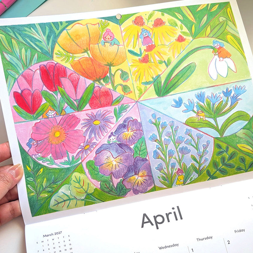 A hand holds a colorful illustrated April calendar page showing floral panels with tulips, daisies, snowdrops, violets, and bluebells above a printed monthly calendar layout.