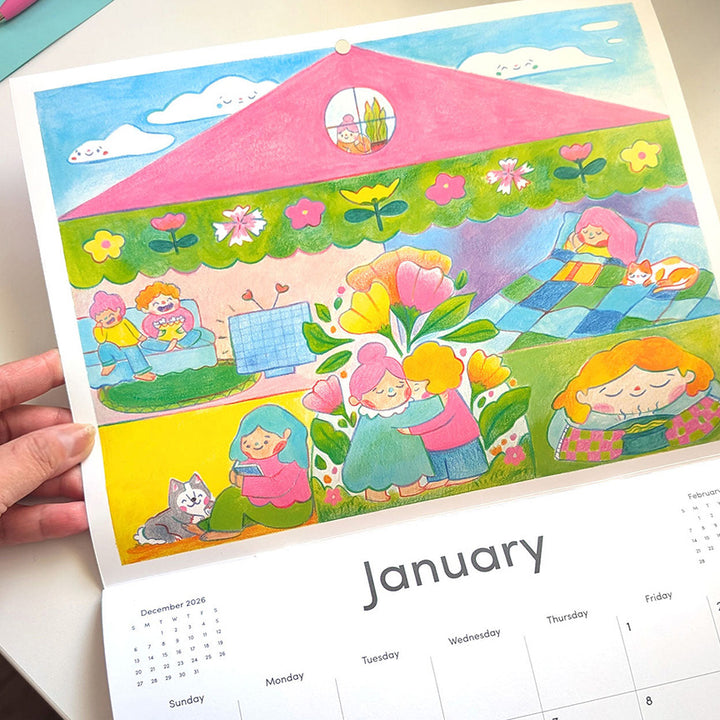 A hand holds a colorful illustrated calendar page showing the inside of a pink house with children, flowers, and a cat, with the word January and a monthly calendar grid visible below.
