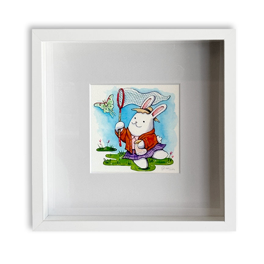 A square white shadow box frame containing a colorful cartoon illustration of a rabbit holding a butterfly net, chasing after a luna moth on a blue sky background.