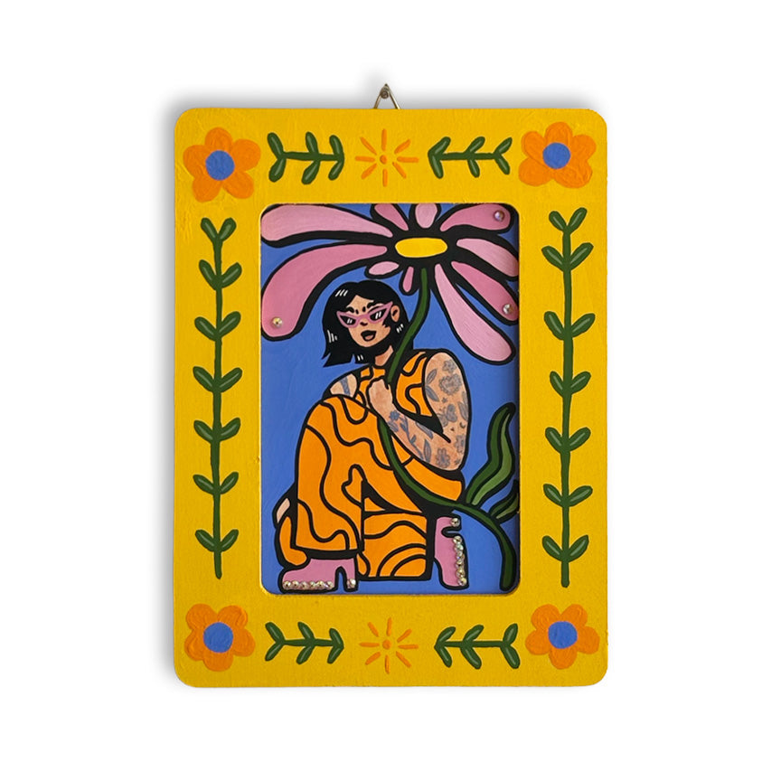 A rectangular art piece with a painted bright yellow frame decorated with orange flowers, green leaves, and small sun motifs. The center contains a colorful illustration of a seated woman holding a large pink flower against a blue background with 3-dimensional gems decorating the flower and her shoes.