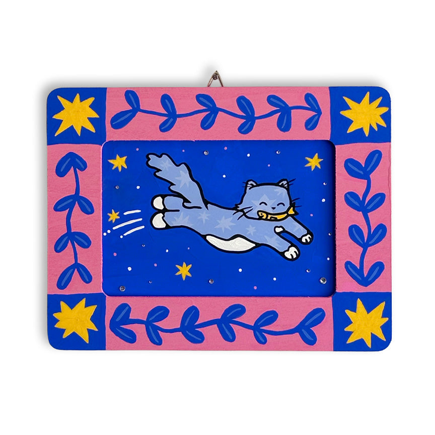 A rectangular art piece featuring a painted blue cat leaping across a starry blue center, surrounded by a painted pink frame with blue leaf motifs and yellow starbursts, shown on a plain white background.