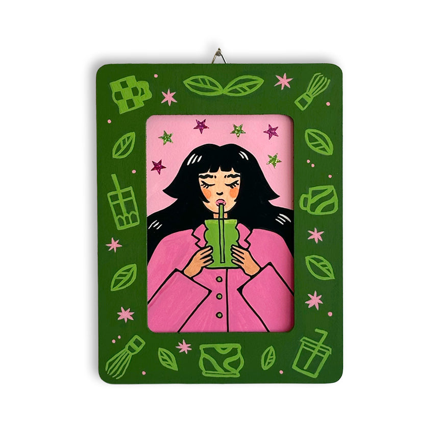 A rectangular art piece with a painted dark green frame and a central pink illustration of a woman drinking matcha. The border includes small light green matcha illustrations and pink stars.