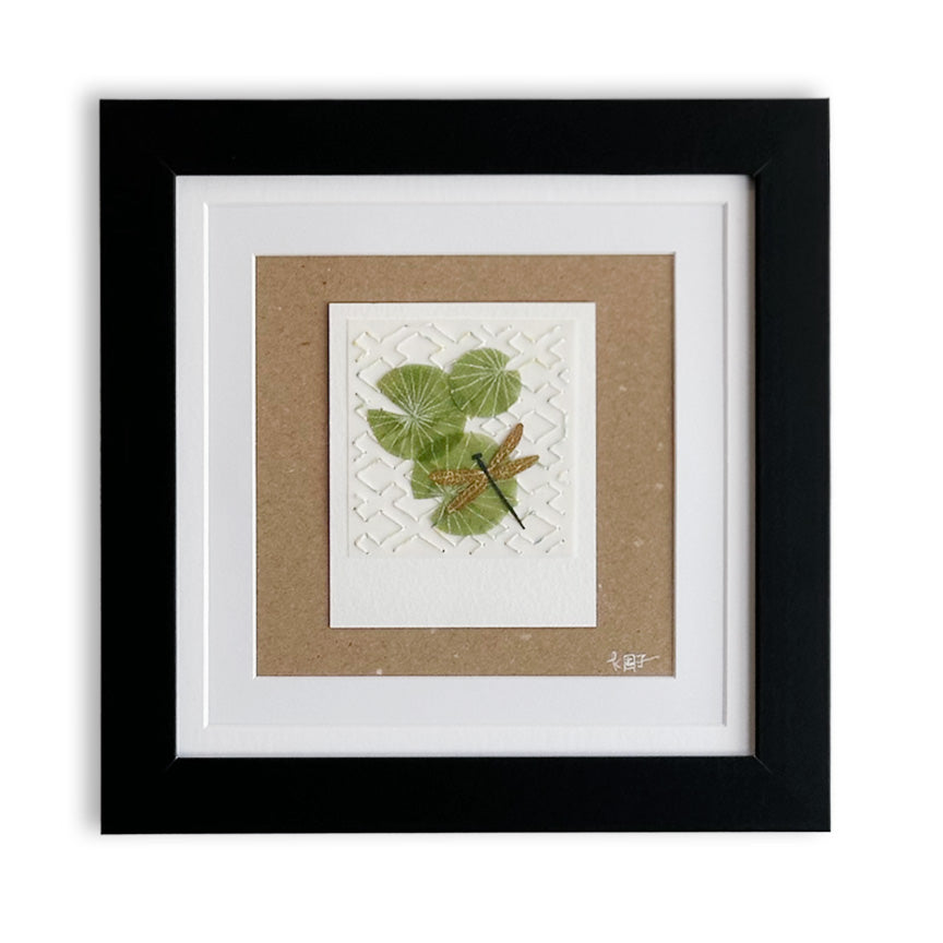 Framed botanical paper cut art piece featuring green lily leaves and a small dragonfly on textured white paper, mounted on a brown paper and set within a black frame with white matting.