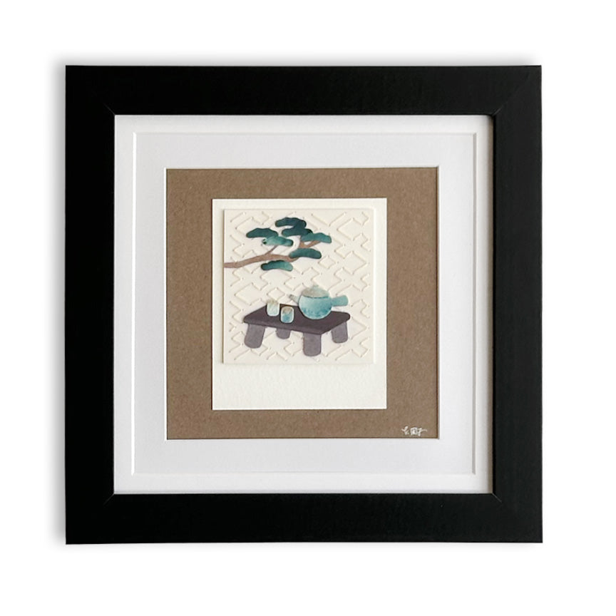 Framed art print featuring a minimalist Japanese-style paper cutting of a small table with a teal cup and a tree branch, set within a black frame with white and tan matting.